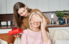 Young woman covering mother's eyes to surprise her with a present in in red wrapping paper at home. 