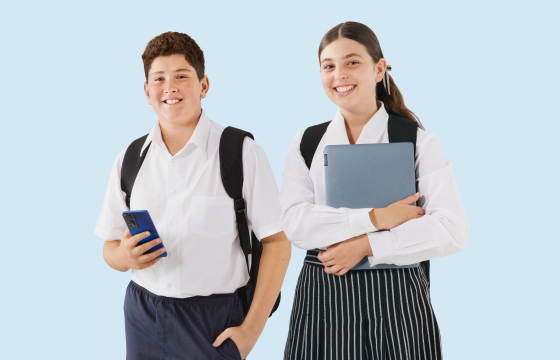 Two high school students in uniforms with backpacks, smiling while holding a smartphone and a laptop against a light blue background.