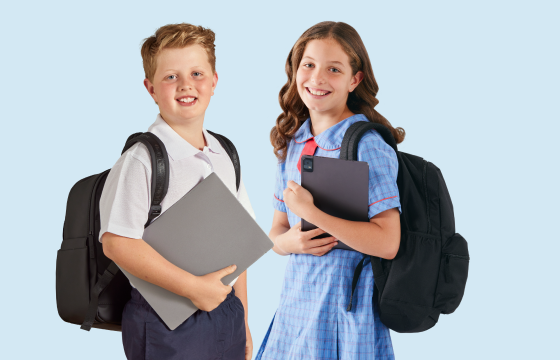 Two primary school students wearing uniforms and backpacks, smiling while holding laptops against a light blue background.