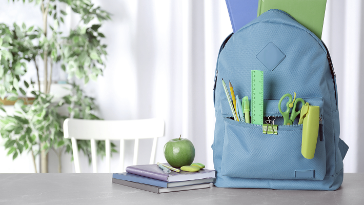 Light blue school backpack filled with stationery items, sitting on a desk beside an apple and stacked notebooks with a plant and chair in the background. 