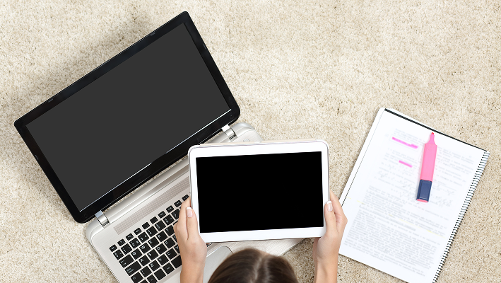 Person sitting on carpet using a tablet in front of a laptop, with a notebook and highlighter beside them. 