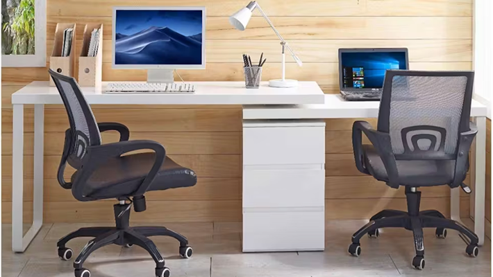 Home office setup featuring two mesh office chairs, a long white desk with desktop and laptop computers, a desk lamp, and wooden wall panelling. 