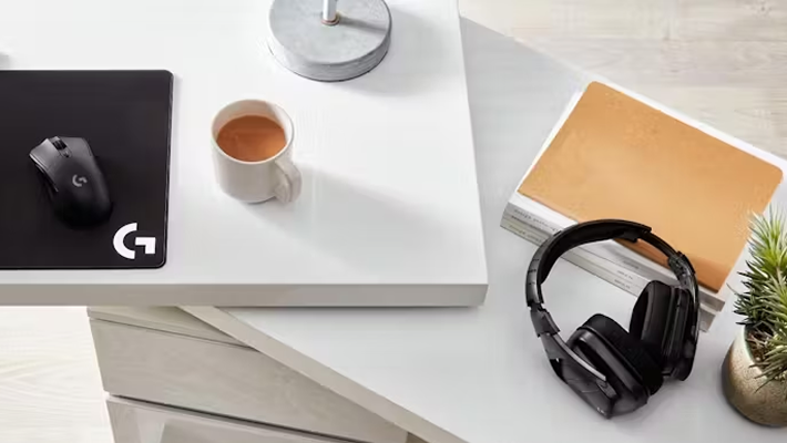 Close-up of a modern white desk with a mousepad, coffee cup, notebook, headphones, and a small potted plant. 