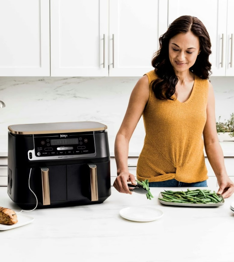 Woman in yellow top preparing vegetables next to a large Ninja dual-basket air fryer in white kitchen