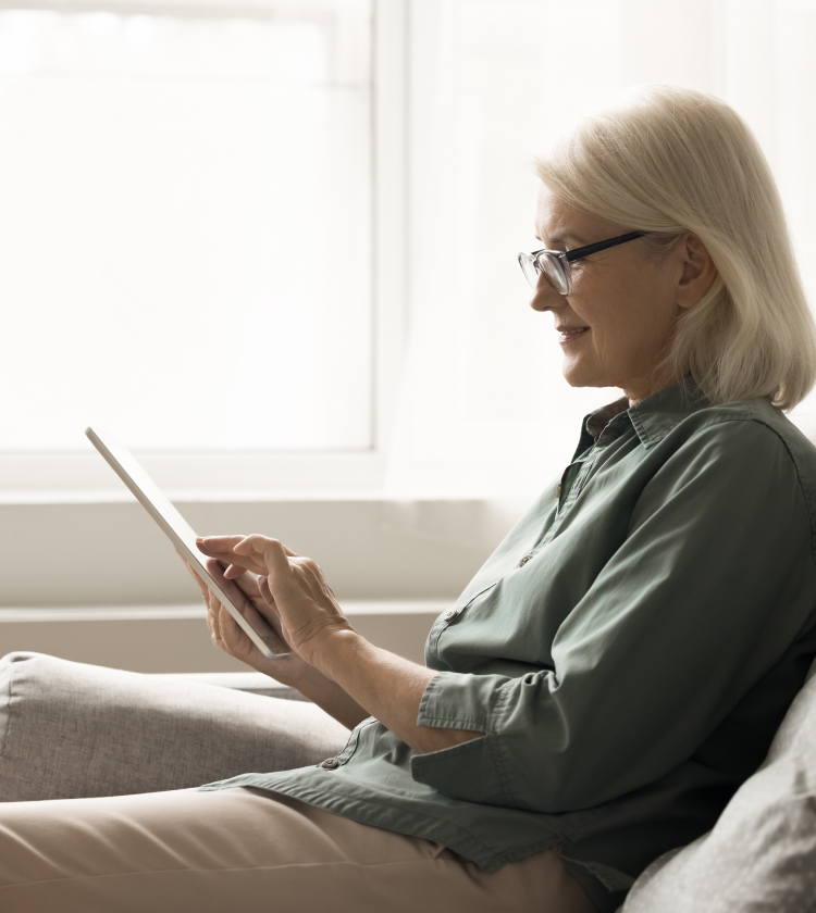 Older woman with glasses relaxing on sofa browsing an e-Reader device at home