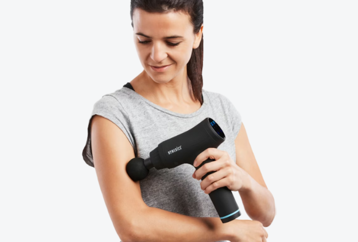 Woman in grey t-shirt using handheld percussion massage gun on upper arm