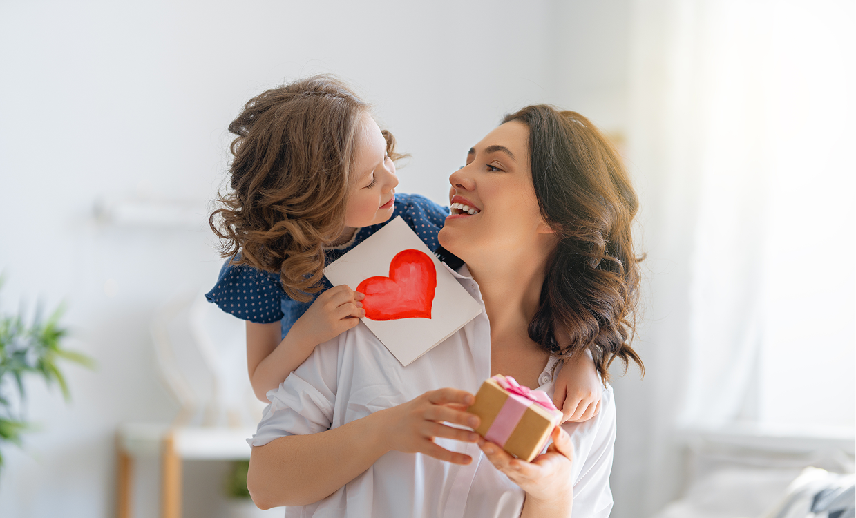 Young child kissing mother on cheek while giving her a handmade heart card and wrapped gift