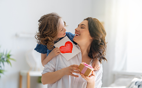 Child leaning in to kiss mother on cheek while holding a handmade red heart card and gift box 
