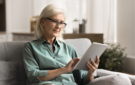 Older woman with glasses sitting on sofa scrolling on a tablet at home. 