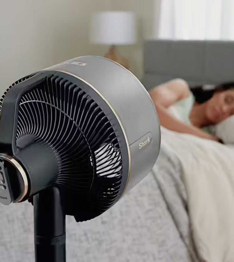 Shark Flexbreeze Pedestal Fan with Misting Attachment positioned beside a bed, with sleeping woman softly blurred in the background 