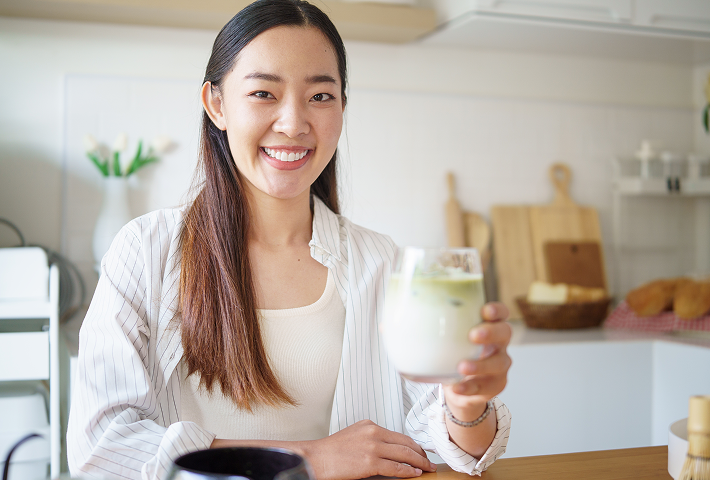 Woman sitting in a bright kitchen holding a creamy frozen drink in a glass. 