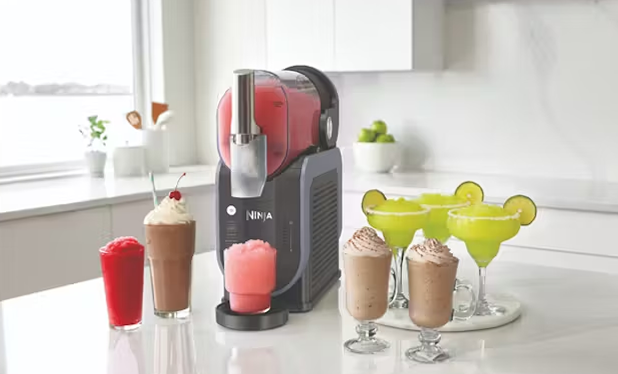 Ninja SLUSHi Professional Frozen Drink Maker on a white kitchen bench, surrounded by a selection of slushies, milkshakes and frozen cocktails in glasses. 