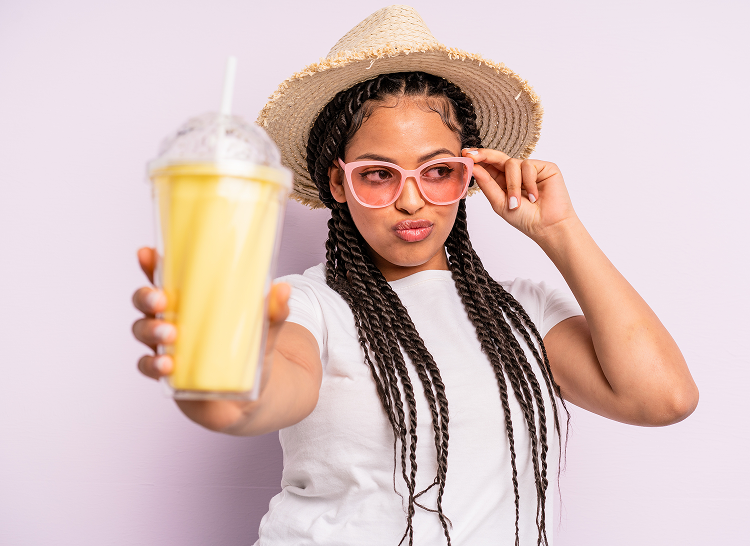 Woman wearing pink sunglasses and a straw hat in front of a light purple background, holding out an orange frozen drink in a clear takeaway cup. 