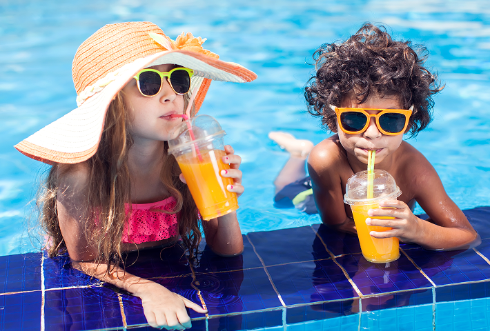 Two children wearing sunglasses, relaxing in a swimming pool while drinking frozen drinks from clear takeaway cups. 