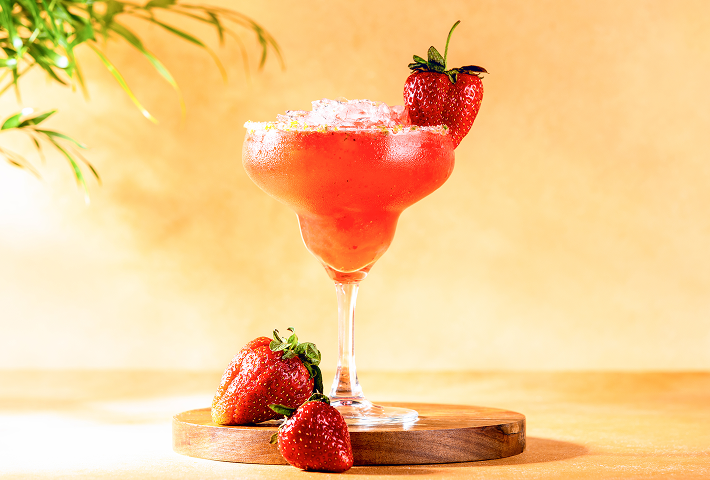 Strawberry frozen cocktail in a margarita glass with a fresh strawberry garnish on a wooden board. 