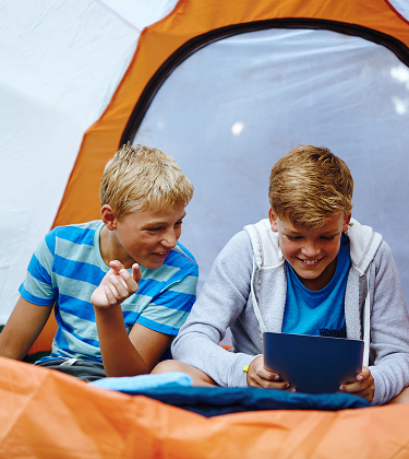 Two young boys sitting inside an orange camping tent using a tablet together 